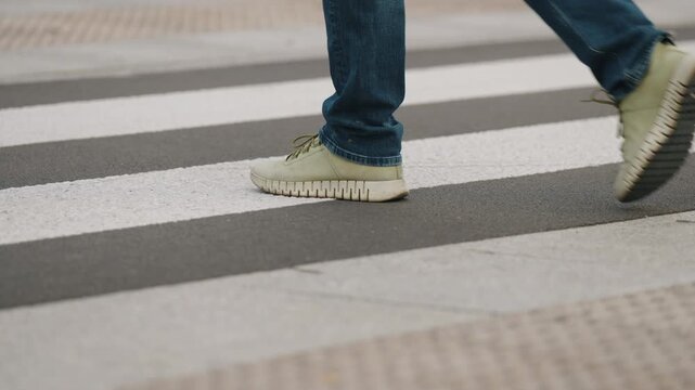 Cinematic close up view urban crosswalk with zebra stripes on asphalt, male pedestrian walking in slow motion, cropped legs to knee level, overcast cloudy city atmosphere