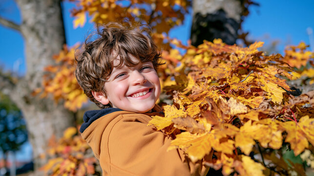 Close up of a smiling young boy hanging playfully from a low tree branch in an autumn park, face framed by golden yellow leaves, tousled hair and warm casual clothing, joyful expression, shallow dep