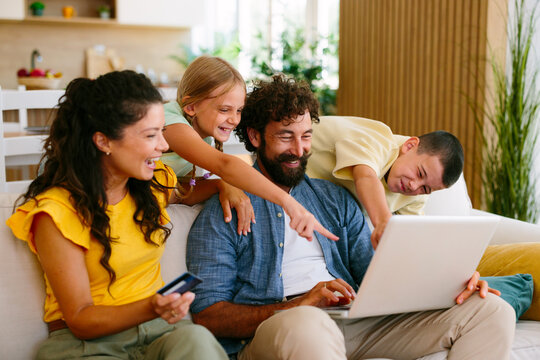 Happy family with children sitting on a sofa at home, enjoying online shopping together, pointing at a laptop screen, and holding a credit card for e-commerce purchases