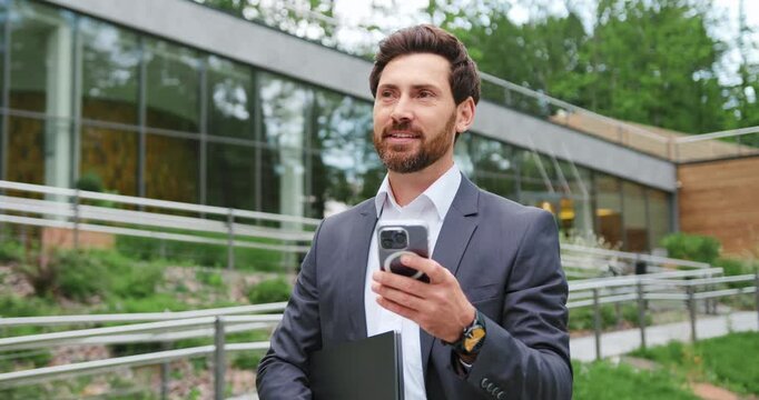 Caucasian bearded businessman using smartphone and holding tablet while looking away outdoors