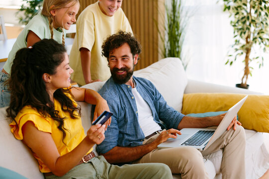 Family sitting on a sofa at home, shopping online together, with the mother holding a credit card and the father using a laptop, children watching happily