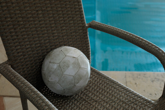 Gray textured exercise ball resting on a woven outdoor chair beside a bright blue swimming pool at a fitness club, this ball sits ready for a poolside ball workout