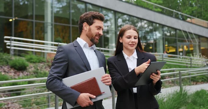 Business man and woman in formal wear using a tablet and discussing work outdoors, representing teamwork, remote work, and professional communication.
