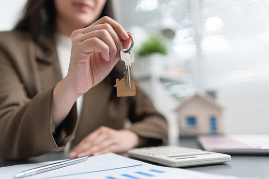 Real estate female agent holds property keys above financial documents, calculator and a model house, representing home ownership
