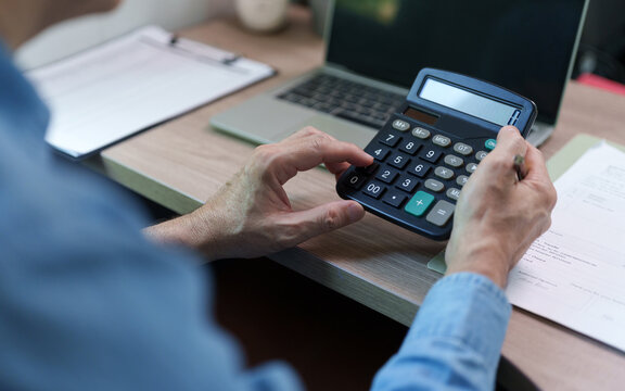 Businessman uses a calculator beside financial documents and a laptop at a desk, representing tax calculation, debt management, budgeting, accounting, financial analysis and planning.
