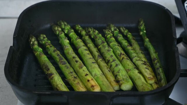 Close up video of freshly washed bright green asparagus spears cooking on a black cast iron griddle pan over a gas stove burner.