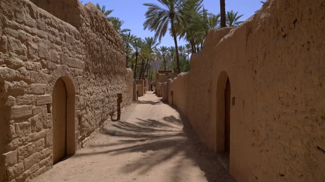 Cinematic shot of sandy heritage walkway between historic mud walls overlooking lush green palm groves in AlUla Old Town