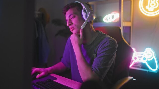 Young man wearing gaming headset yawns with hand to mouth at computer desk in studio with neon controller sign; late night fatigue.