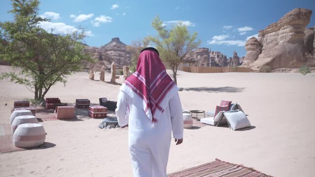 Cinematic back view of Saudi man in traditional thobe and shemagh walking around luxury desert resort courtyard in AlUla