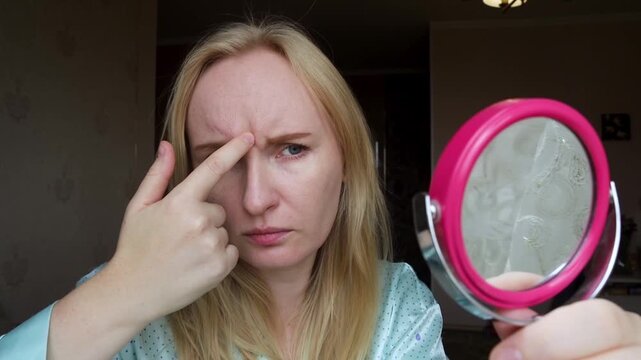 Concerned adult woman looking in hand mirror, examining forehead wrinkles and glabellar frown lines. She touches her skin, worrying about aging, skincare, and botox injections.