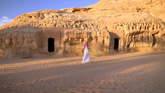 Cinematic wide shot of Saudi man walking on sand past a massive sandstone mountain cliff with row of carved tombs in Madain Saleh AlUla