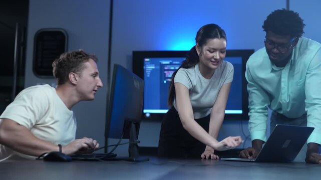 Asian female and African American male software developers discussing code on laptops with Caucasian colleague. Diverse tech team collaborating in modern IT office. Corporate technology, business data