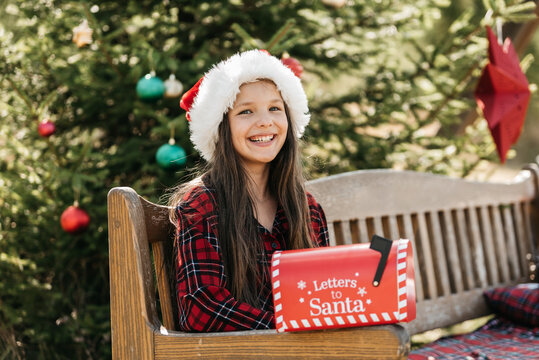Christmas in July with girl wearing Santa hat and holding red Santa mailbox outdoors near decorated Christmas tree in summer garden. Christmas and New Year traditions to order gifts by mail from Santa