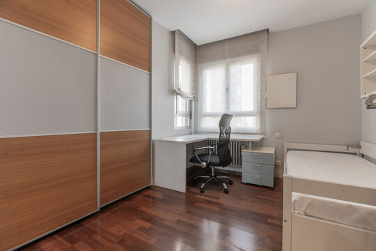 Bedroom with two-tone wardrobe, corner desk and office chair, small dresser, and daybed near a curtained window.