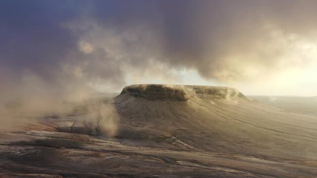 inematic aerial drone shot of dense clouds fog and light snowfall wrapping around volcanic plateau mountain peak at sunrise in Harrat Tabuk
