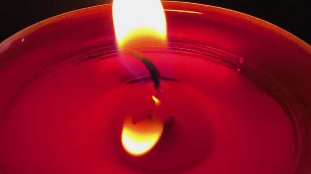 Close-up of a burning red tealight candle with a bright flickering flame reflected in melted wax, creating warm dramatic illumination, romantic atmosphere, relaxation, and peaceful meditation concept.