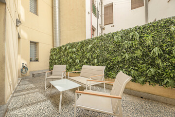 Fototapeta premium Sunlit inner courtyard with lounge chairs, sofa, and lush living plant wall between building facades.