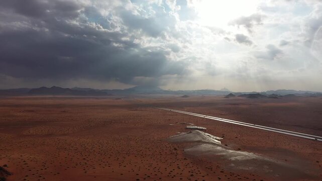 Cinematic aerial drone shot of long straight desert highway across red sand during rain shower with sunbeams breaking through clouds in Saudi Arabia