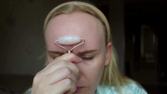 Concerned woman uses a quartz facial roller to massage forehead wrinkles and glabellar lines. She looks in the mirror, seeking anti aging skin care alternatives to botox.