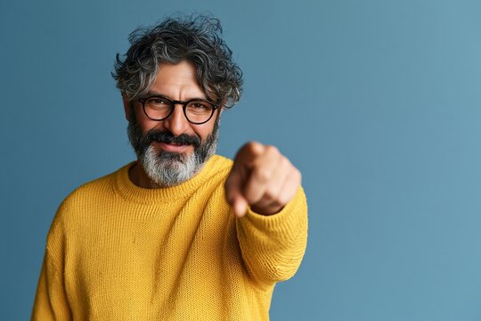Middle aged man with curly hair and beard wearing yellow sweater pointing forward at camera