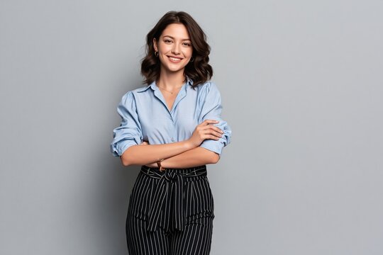 Smiling young woman with dark wavy hair wearing blue shirt and patterned trousers against gray