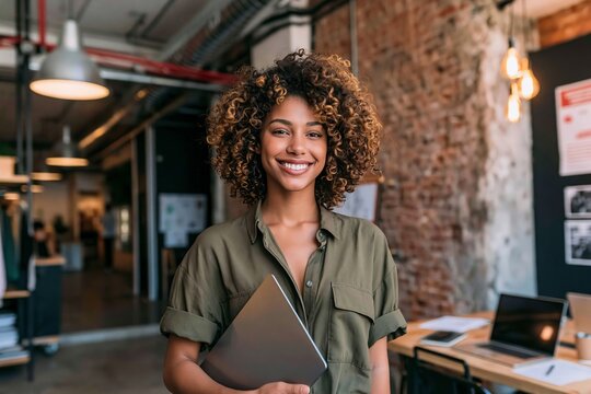 Smiling young woman with curly hair holding laptop standing in modern creative office interior