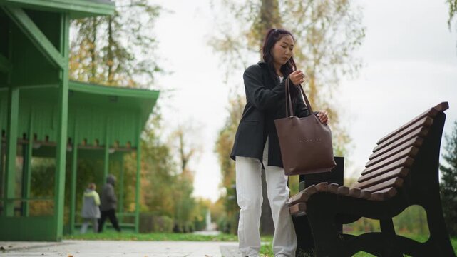 Close up of young woman walking through outdoor park carrying brown handbag and wearing white pants. Woman approaches camera with trees in autumn foliage visible behind her. scene includes