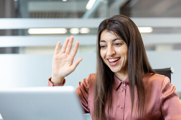Fototapeta premium Professional young woman engaging in a virtual meeting, smiling and waving a hand while communicating remotely from a modern office environment using her laptop