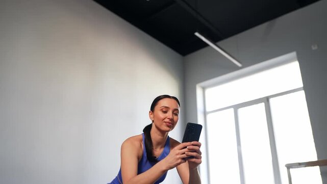Portrait of charming active fit woman in sportswear making video call, communicating online with friend using mobile phone, sharing about workout routine while exercising squats in fitness studio.