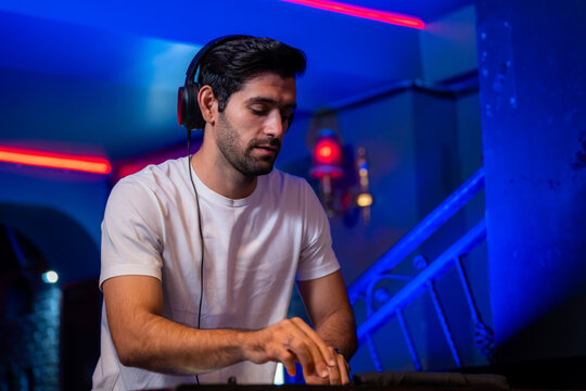 A young bearded male DJ in a white t-shirt working the console of a neon-lit club keeps the beat with a small nod of his head at the DJ booth of a neon-lit nightclub.