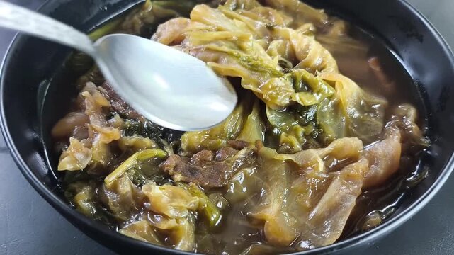 Close-up of a metal spoon scooping traditional Thai-Chinese mixed vegetable stew (Tom Jab Chai) from a black bowl. 