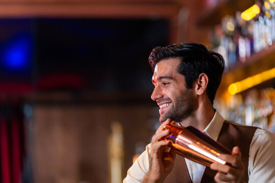 A smiling male bartender in a brown waistcoat shaking a copper cocktail shaker behind the bar holds the copper shaker up in front of his chest behind the counter of a warm lit cocktail bar.