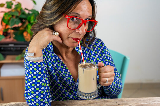 Woman in patterned shirt enjoying iced coffee with a straw, wearing red glasses