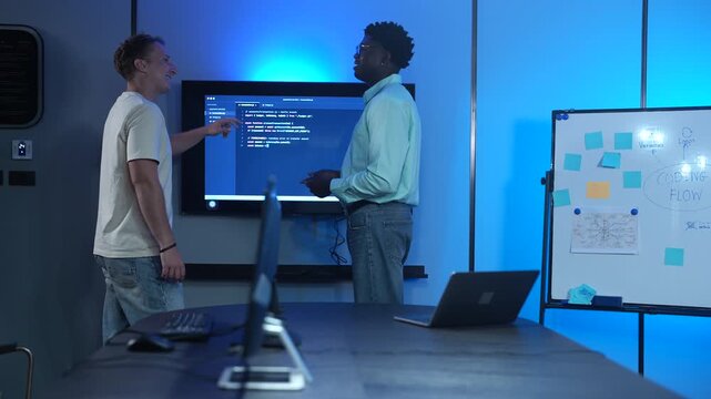 African American IT manager giving technical briefing to Caucasian developer in modern offices. Diverse male programmers discussing software presentation on screen. Corporate technology teamwork 