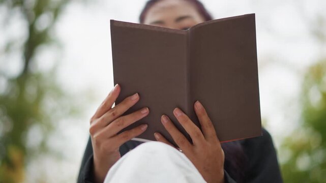 Close up of young woman holding and reading open book outdoors. Her hands are visible gently grasping brown cover while woman focuses on its pages. background is softly blurred with natural