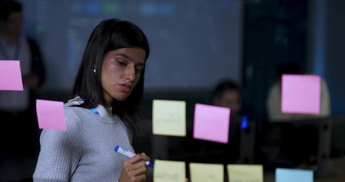 Adult female technology professional writes on sticky notes at glass board, reviewing project tasks and organizing software workflow during collaborative strategy planning in modern corporate office.