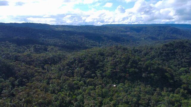 Wide aerial drone view of endless forested hills rolling toward a distant plateau in the Amazon foothills of Ecuador under a bright sky with scattered clouds.