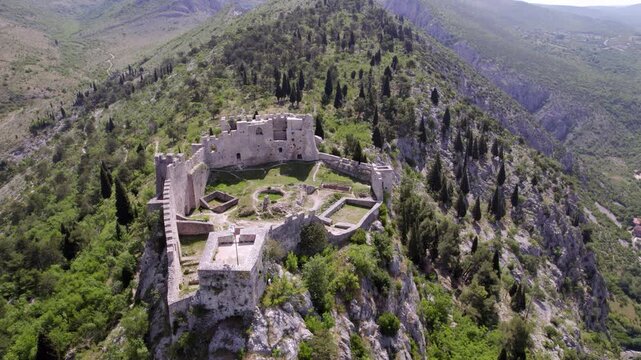 Blagaj Fortress crowns a rocky ridge surrounded by mountain woodland in Bosnia and Herzegovina, aerial high establishing orbit overview