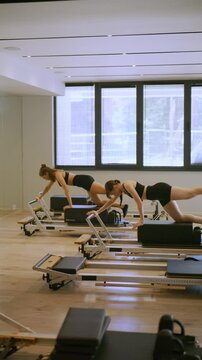 Two young women are performing exercises on Pilates reformers in a bright studio. They are engaged in a fitness workout, demonstrating strength and flexibility.