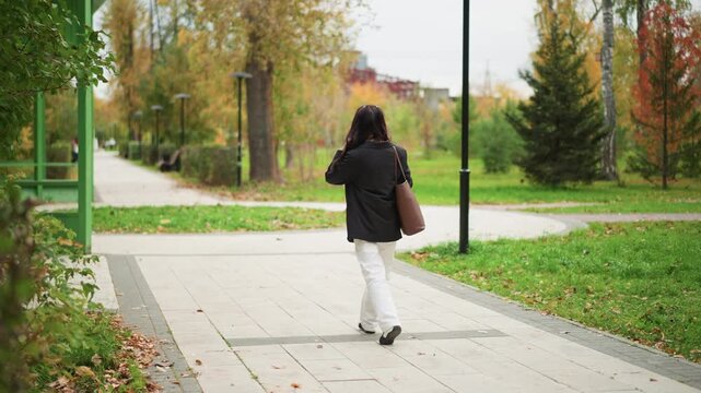 Woman with long dark hair walks away from camera along paved path in outdoor park carrying brown shoulder bag. Woman wears black coat and white pants as woman moves steadily forward