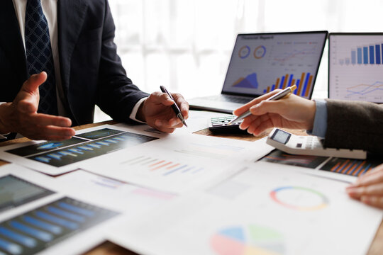 Business team collaborating around laptops and printed reports, analyzing charts and financial data, strategizing growth, performance and planning in a modern office setting