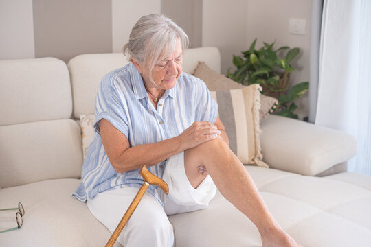 Elderly woman suffering from intense knee pain, sitting on a sofa with her walking stick nearby, highlighting health challenges and the need for support in daily life