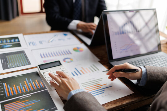 Business professionals analyzing financial data and statistics, using a calculator and charts on paper, with a laptop displaying graphs, planning company growth and strategy