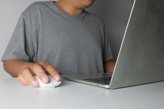 Close up of a man in a casual grey shirt using a white wireless mouse and laptop on a desk. Concept of working from home, freelance, online business, education, and internet surfing.