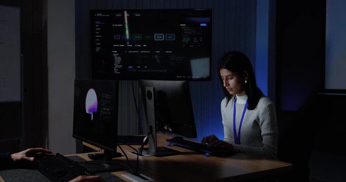 Adult female technology professional types at computer as adult male colleague approaches, leans beside workstation, and joins software dashboard review during collaboration inside modern office.