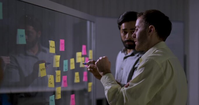 Two adult male technology professionals discuss sticky notes on glass board, pointing at project ideas and reviewing workflow during collaborative software strategy planning session in modern office.
