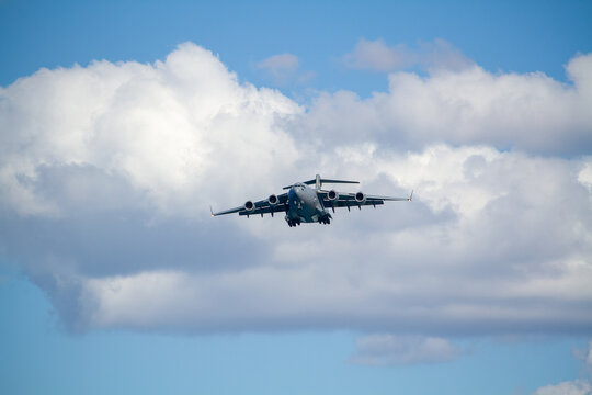 C-17 Globemaster III military transport aircraft in flight, viewed from below against a blue sky with a white cloud below the aircraft, showing its four jet engines and distinctive high-wing design.