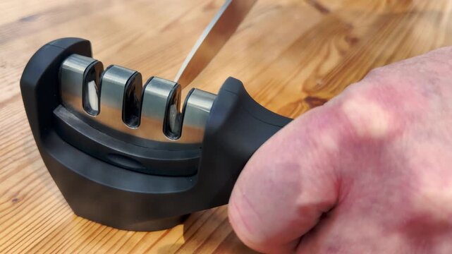 A man&rsquo;s hand pulling the steel blade of a large kitchen knife through a modern ergonomic 3 stage sharpener, while the other hand holds the tool steady.