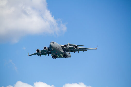 C-17 Globemaster III military transport aircraft in flight, viewed from below against a blue sky with a white cloud below the aircraft, showing its four jet engines and distinctive high-wing design.