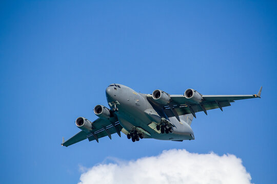 C-17 Globemaster III military transport aircraft in flight, viewed from below against a blue sky with a white cloud below the aircraft, showing its four jet engines and distinctive high-wing design.